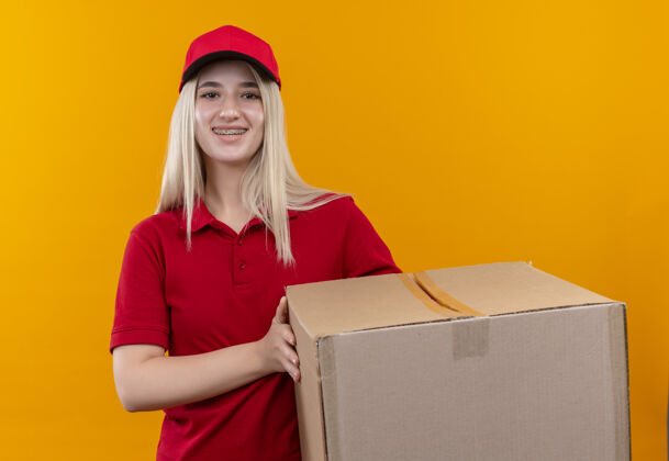 支撑微笑的小女孩穿着红色t恤 戴着帽子 戴着牙套 在孤立的橙色背景下拿着盒子帽子盒子橙色
