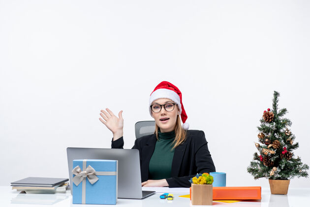 坐着一位年轻漂亮的女士戴着圣诞老人的帽子坐在一张桌子旁 桌上放着圣诞树和礼物 谈论着办公室里的一些事情笔记本电脑圣诞老人有魅力的女人