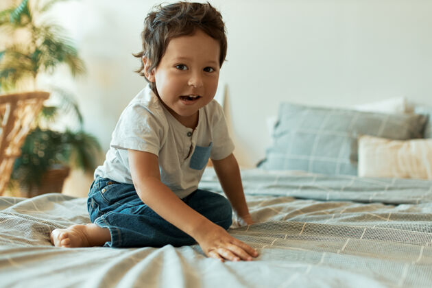 欢快室内肖像可爱的婴儿深色皮肤 卷发和赤脚坐在床上的t恤和牛仔裤早晨儿子混合