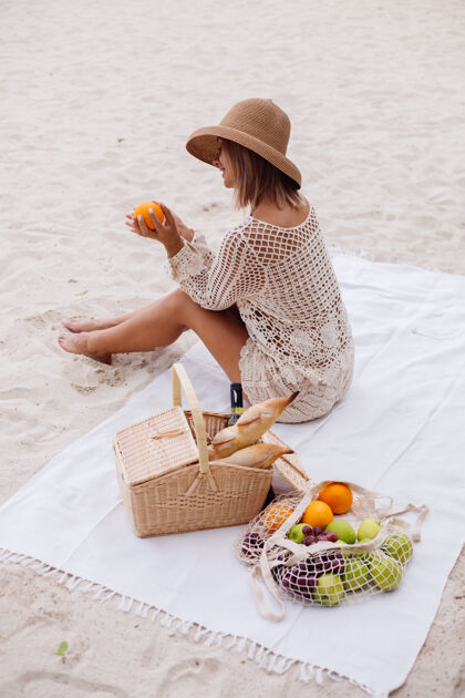 水果一个年轻的女人坐在毛巾上 戴着草帽 穿着白色针织衣服和野餐篮子野餐浪漫热带