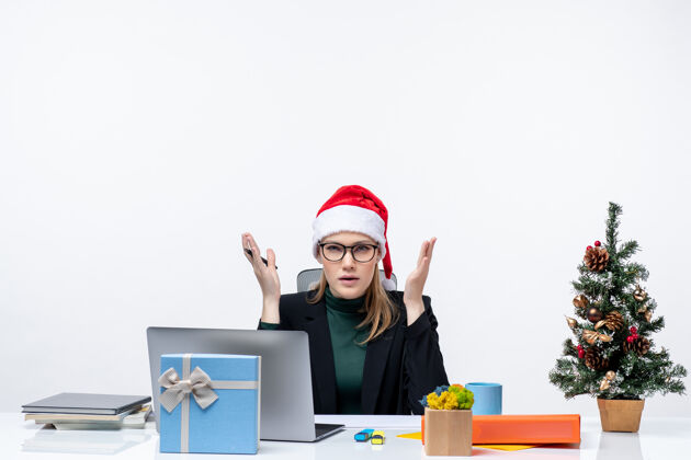 礼物一个困惑的金发女人 戴着圣诞老人的帽子 坐在一张桌子旁 桌子上放着一棵圣诞树和一份礼物 在白色背景下质疑办公室里的一些东西办公室电脑女人