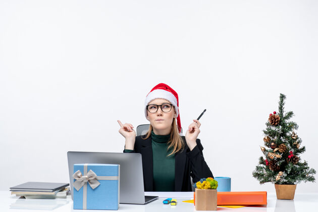 电脑困惑的金发女人 戴着圣诞老人的帽子 坐在一张桌子旁 桌子上放着一棵圣诞树和一份白色背景的礼物办公室坐着圣诞节