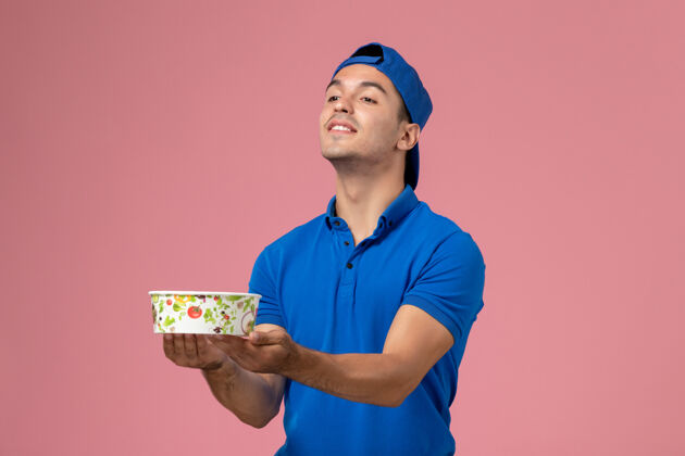 视图正面图身穿蓝色制服披肩的年轻男性信使手持浅粉色墙上的圆形投递碗递送人年轻男性信使