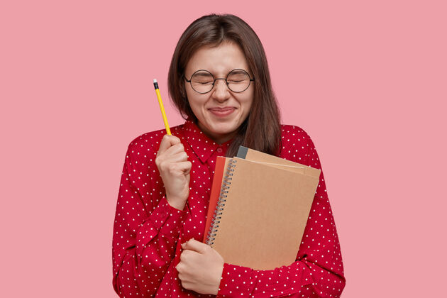 衬衫充满希望的女学生闭上眼睛 手里拿着铅笔 拿着记事本 相信能听到考试结果 戴着圆眼镜和红衬衫 隔着粉色的工作室墙装备学生姿态