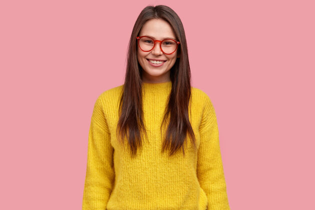 工作满意的漂亮黑发年轻女子的上身镜头 表情愉快 戴着眼镜和黄色休闲套头衫 粉色背景下的模特成功满意眼镜