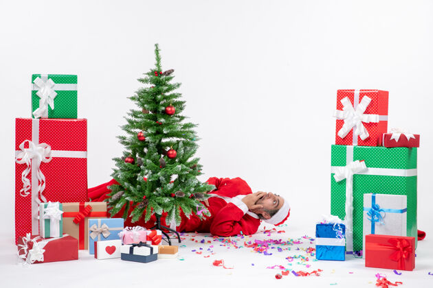 圣诞节日气氛与年轻的惊喜圣诞老人躺在圣诞树后靠近礼物白色背景冬青节日惊喜
