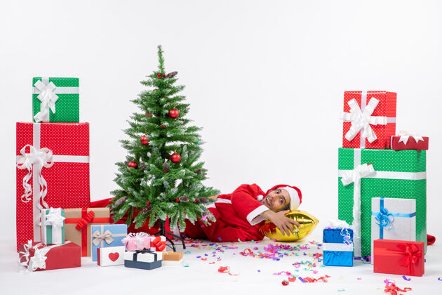 谎言圣诞老人躺在圣诞树后面 在白色背景画面上不同颜色的礼物旁边 充满节日气氛冬青节日礼物