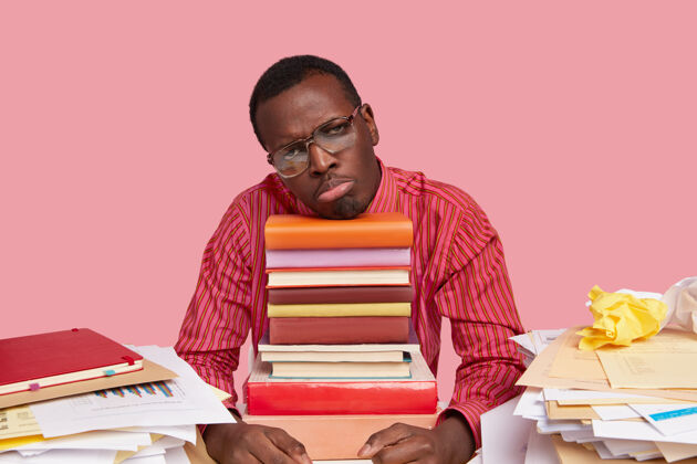 家庭作业不快乐的黑皮肤男人穿着正式的衣服 失望 厌倦了工作 倚在一堆书上 戴着大眼镜和正式的衬衫眼镜书桌学生