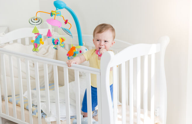 卧室伤心的10个月大的男婴哭着抱在婴儿床旁边婴儿床就寝时间护理