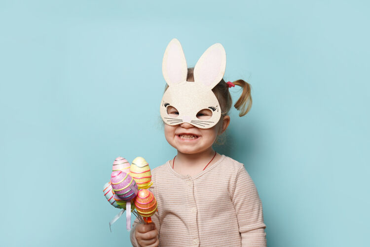玩一个可爱的小女孩穿着复活节兔子耳朵拿着彩蛋在蓝色表面的肖像笑季节节日