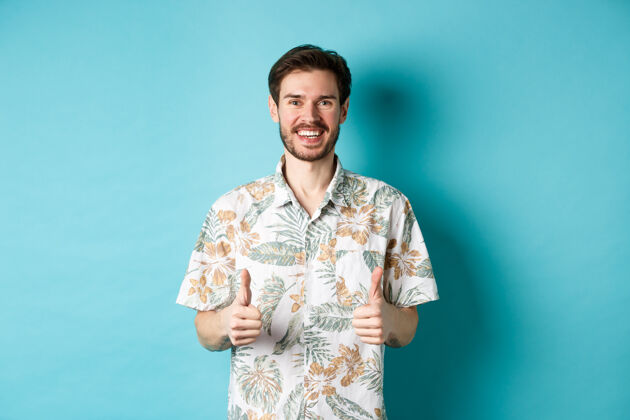 年轻暑假快乐的游客竖起大拇指 微笑着 穿着夏威夷衬衫 推荐旅行社 蓝色背景站休闲成年人