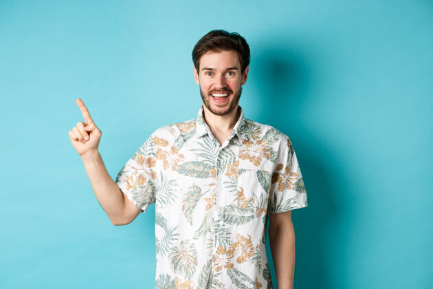 男性一个穿着夏威夷衬衫的快乐的年轻人用手指着左边 露出空荡荡的笑容旅游度假的概念夏威夷人年轻站着