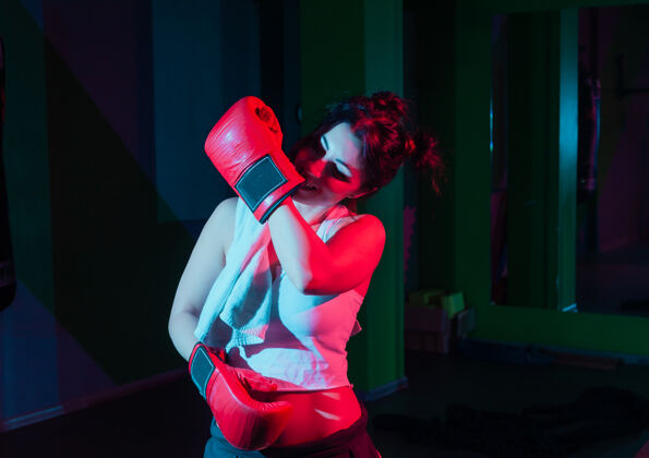 战斗年轻女子拳击手戴着拳击手套 肩上扛着毛巾 在黑暗的墙上霓虹灯渐变着红蓝色的灯光教练女子运动服