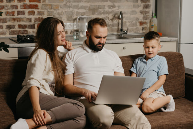 人一位留着胡须的父亲正在远程操作笔记本电脑 而他的儿子和妻子则在盯着屏幕沙发女孩女性
