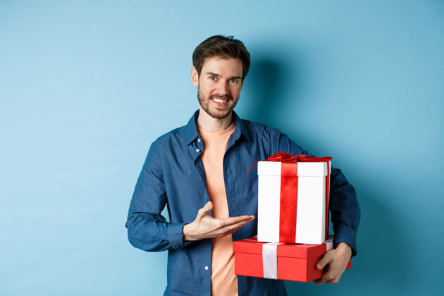 微笑穿着休闲服的年轻人在情人节买浪漫的礼物 指着礼物盒微笑着 站在蓝色的背景上温柔激情惊喜