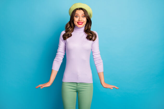 情绪照片中惊人的旅行者女士笑容可掬时尚造型好心情穿现代绿色贝雷帽紫色高领套头衫裤子隔离蓝色墙壁休闲欢乐高领毛衣