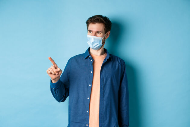 隔离英俊的年轻人戴着冠状病毒的医用口罩 指着左边的手指 看上去很满意 微笑着看着空旷的空间 蓝色的背景医疗男人朋友