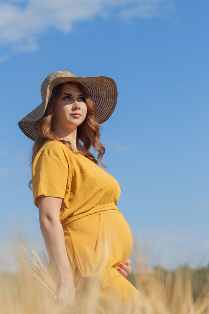 阳光在一个阳光明媚的夏日 一位年轻漂亮的孕妇穿着黄色的裙子 戴着黄色的帽子 走过一片麦田欢呼太阳人