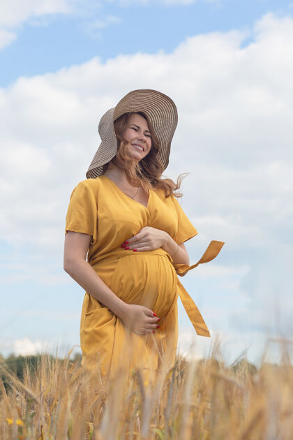 太阳在一个阳光明媚的夏日 一位年轻漂亮的孕妇穿着黄色的裙子 戴着黄色的帽子 走过一片麦田阳光可爱天空