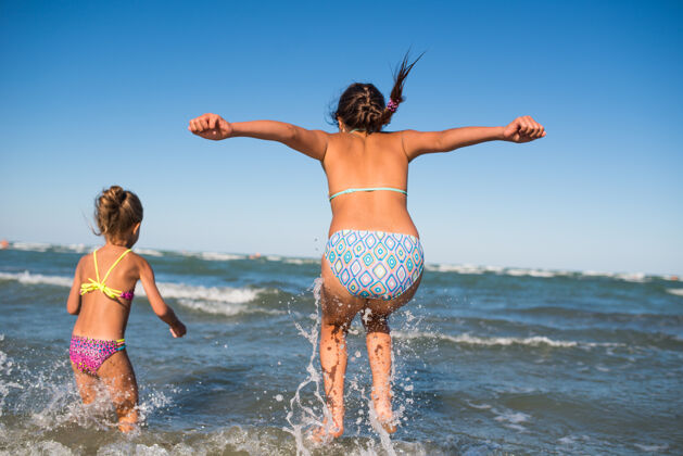 海滩两个有趣的小女孩在喧闹的海浪中跳跃 在一个阳光明媚的夏日享受期待已久的假期跳跃无云风
