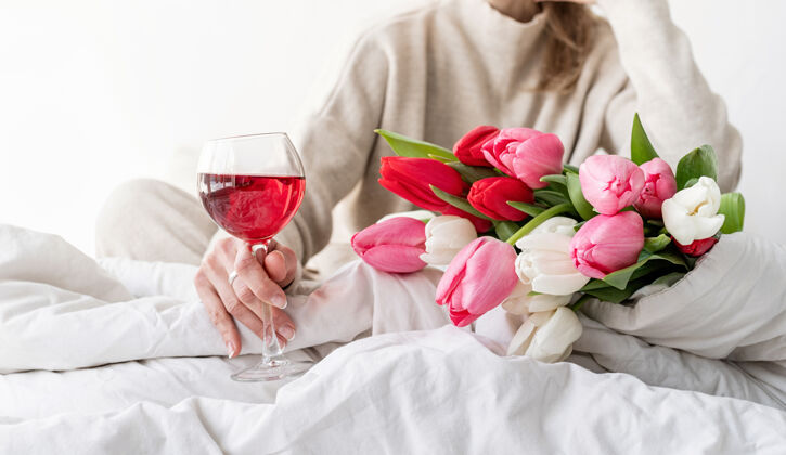 节日快乐的女人穿着睡衣坐在床上 愉快地欣赏着鲜花和一杯红酒妈妈情人节情绪