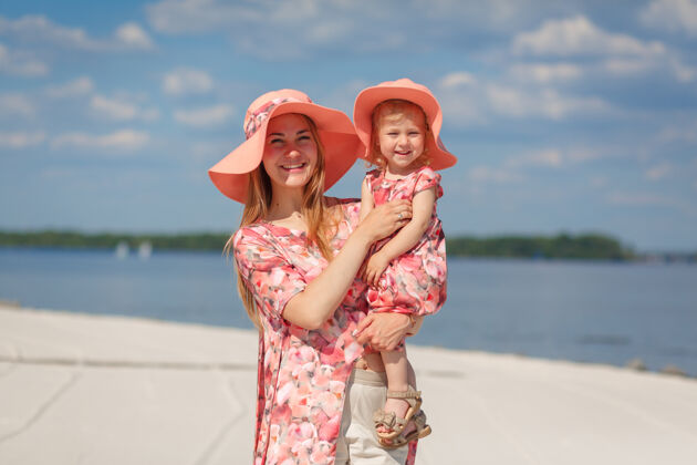 母亲一个穿着夏日太阳裙的迷人女孩和她的小女儿在沙滩上散步享受温暖晴朗的夏日笑帽子衣服