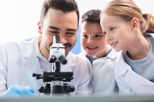 女孩期望结果令人愉快当孩子们看着他咧嘴笑的时候 快乐的男人在用显微镜实验室知识面子