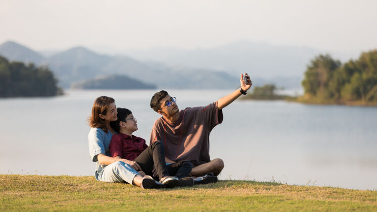 泰国亚洲人一家三口 母亲和两个年幼的儿子 一起坐在一个有山有水的巨大湖边背景他们用智能手机拍照电话妈妈夏天