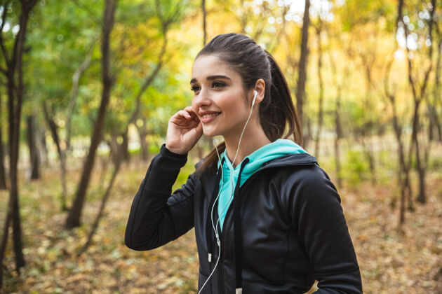 减肥微笑的健身女 戴着耳机听音乐 手持手机站在公园里户外公园森林