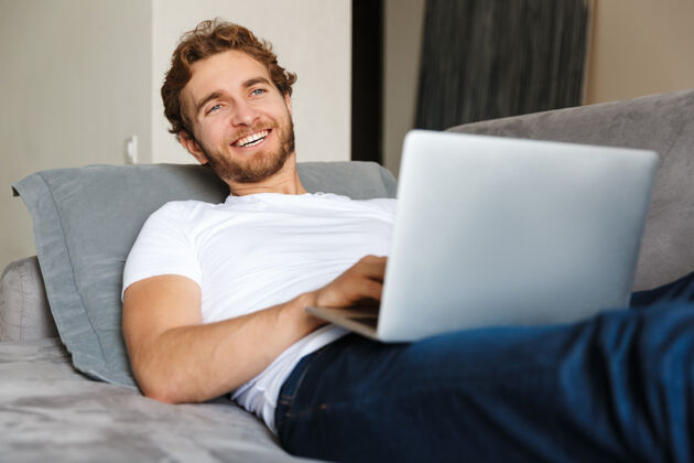 学习一个留着胡子的帅哥坐在家里的沙发上用笔记本电脑吸引休闲舒适