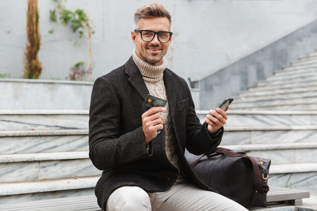 信用帅哥穿着夹克坐在户外用手机和信用卡上网购物户外移动户外