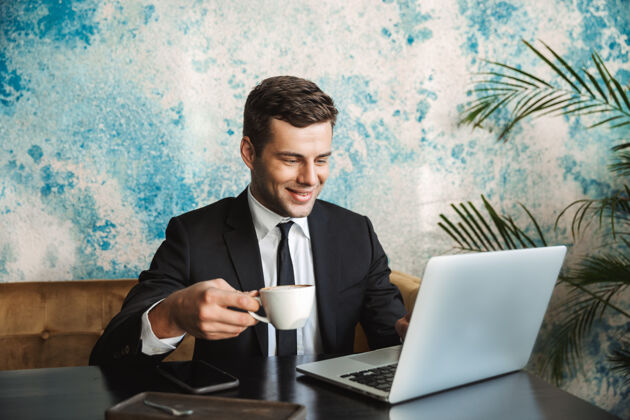 个人电脑一个英俊快乐的年轻商人坐在咖啡馆里用笔记本电脑喝咖啡的画面企业家网络笔记本电脑