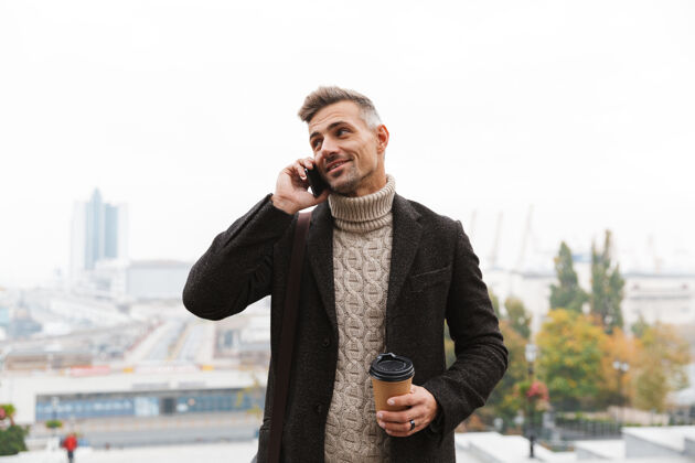 城市30多岁的帅哥穿着夹克 手持手机 端着外卖咖啡 走在城市街道上杯子手机成人