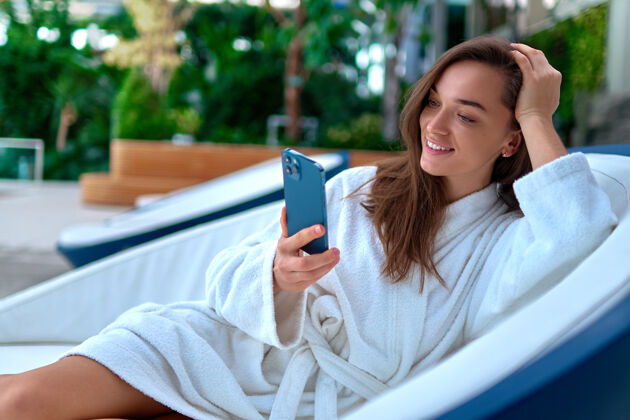 度假村穿着白色浴袍的年轻迷人的微笑女子 在温泉度假区放松时躺在躺椅上 使用智能手机观看视频和在线浏览休息聊天白天