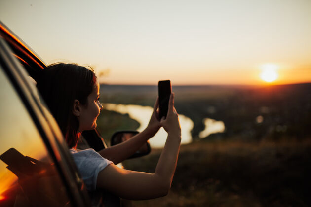 周末年轻的女旅客在车内 看着美丽的日落 拍照留念享受假日女孩