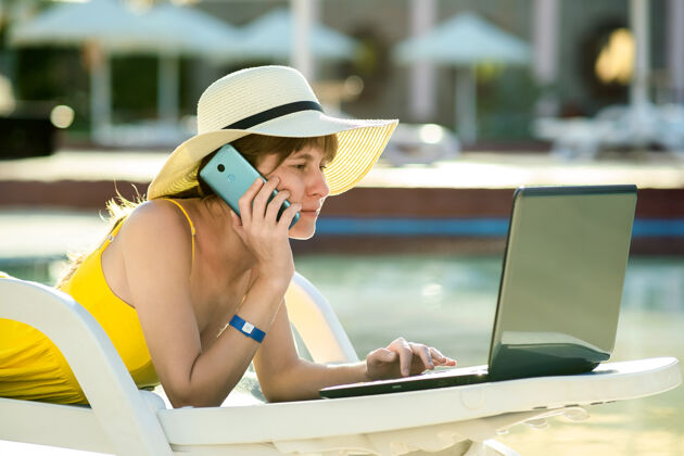 网站在避暑胜地 一个年轻的女人坐在游泳池的沙滩椅上 用笔记本电脑和电话交谈女人在线游客