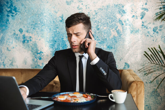 企业家一个英俊的年轻商人坐在咖啡馆里用笔记本电脑吃早餐或晚餐 通过电话交谈的画面帅气饮食男士