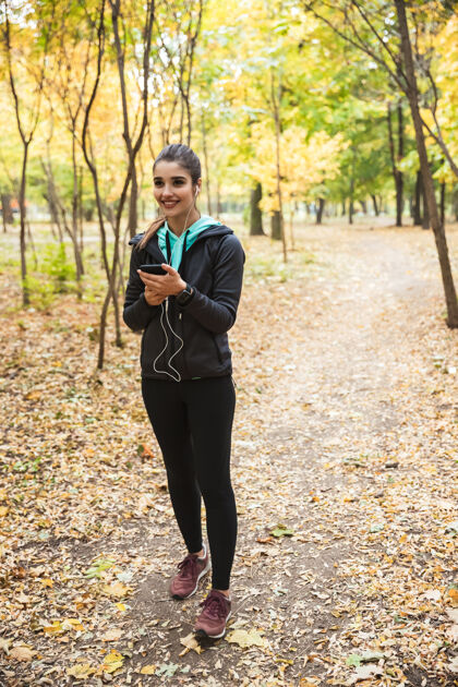 花园照片中 一位年轻漂亮的健身女士在公园里用手机和耳机听音乐姿势移动放松