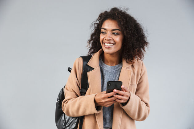 电话微笑的非洲年轻女子穿着外套 一边用手机 一边孤立地站着肖像眼镜夹克