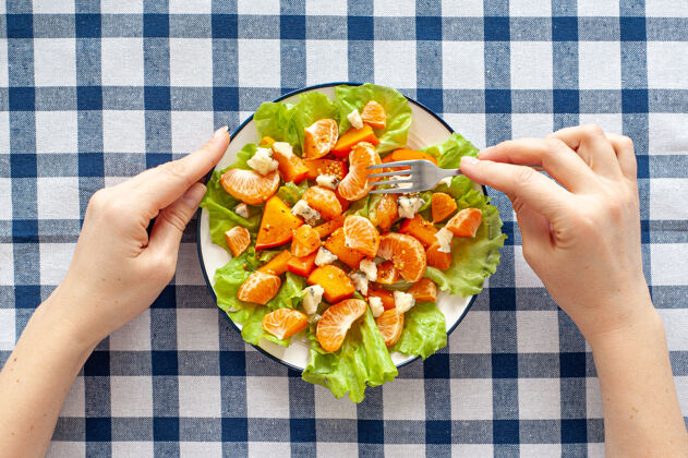 素食明亮的维生素沙拉 柿子 橘子和蓝奶酪在格子桌布上女人的手拿着叉子和一盘沙拉膳食维生素食物从上面看盘子饮食食物