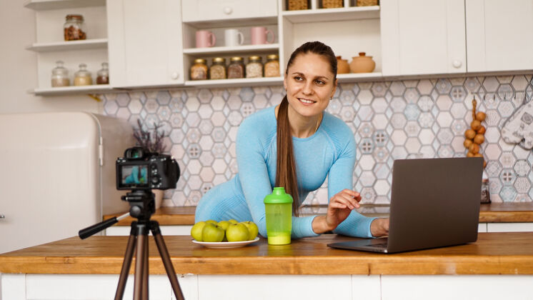 笔记本厨房里拿着笔记本电脑的年轻女子微笑着美食博客的概念一位女士正在录制一段关于健康饮食的视频三脚架上的相机女人厨房拍摄
