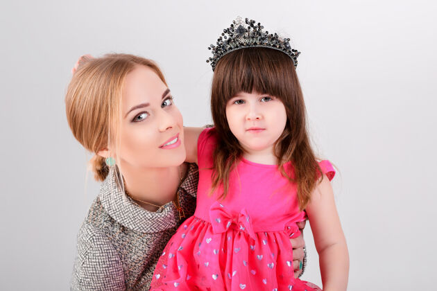 童年美丽的小女孩穿着粉色公主裙 头戴皇冠 美丽的母亲在灰色背景下拥抱着婴儿欢呼漂亮