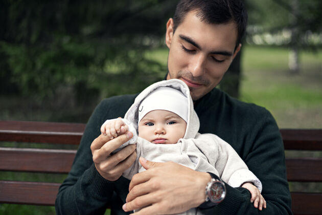 认证特写年轻的父亲穿着绿色套头衫 怀里抱着孩子坐在公园的长凳上男人家庭童年