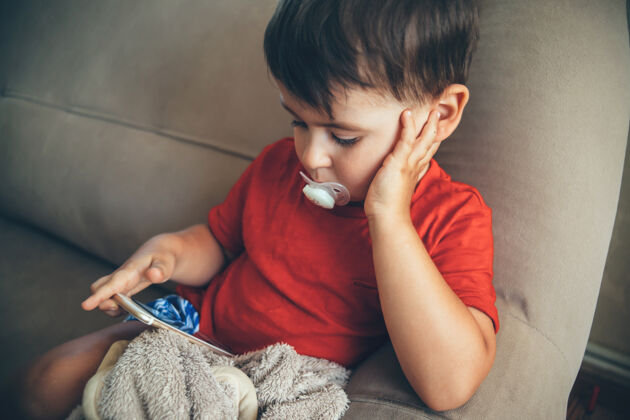 黑发一个白人小男孩坐在床上看着手机屏幕 嘴里叼着喂食器假日享受微笑
