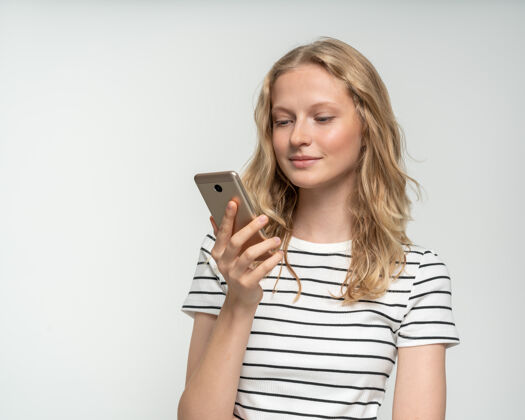 打字站在白墙附近 灰色背景下 漂亮的女性在手机上查看社交媒体长发金发聊天