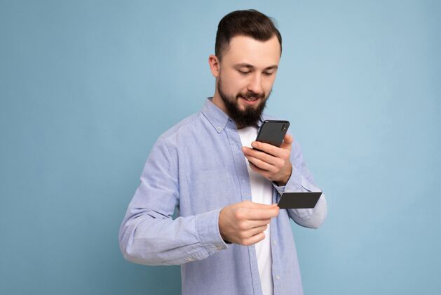 蓝色背景帅哥穿着日常的衣服 隔着背景墙拿着手机和信用卡付款 看着智能手机屏幕 好看年轻男性