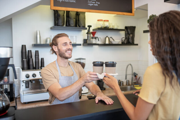 咖啡师咖啡去吧礼貌点站在咖啡店柜台后面的年轻的大胡子咖啡师给长发女人两杯咖啡腰向上订单咖啡店