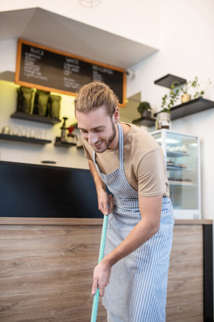 咖啡馆顺序和清洁 细心微笑的男人穿着条纹围裙 拿着拖把在咖啡馆的吧台上打扫卫生心情好看湿洗