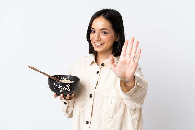 日本人年轻的白人妇女被隔离在白墙上 一边用筷子端着一碗面条 一边高兴地用手敬礼你好再见黑发