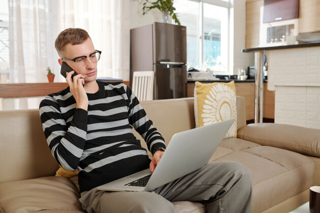 打字戴眼镜的严肃的年轻商人坐在家里的沙发上 打电话 在笔记本电脑屏幕上阅读文件休闲活动休闲服年轻人